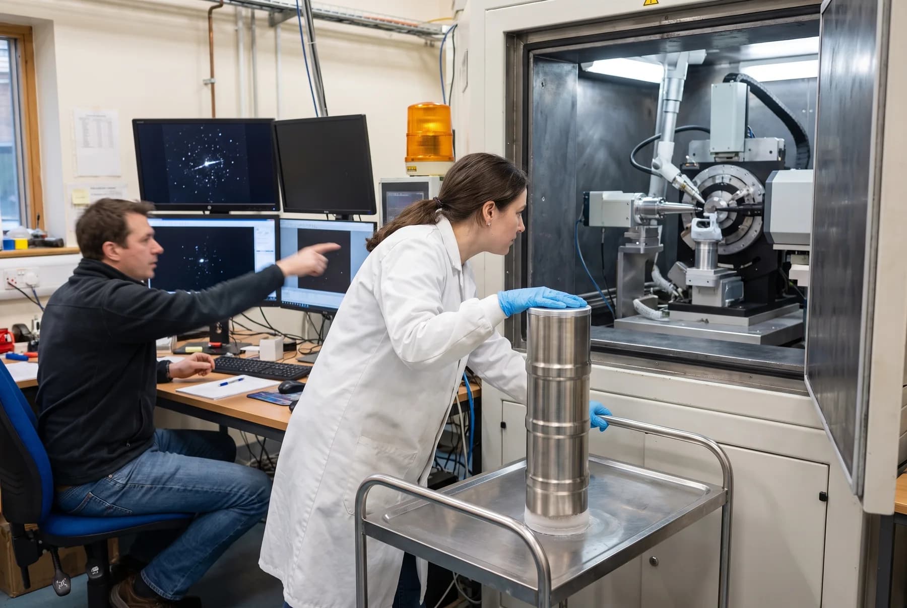 A researcher in a lab coat and blue gloves resting a hand on Crìa Go LN₂ on a cart in front of an X-ray diffractometer, a colleague pointing at a diffraction pattern on screen